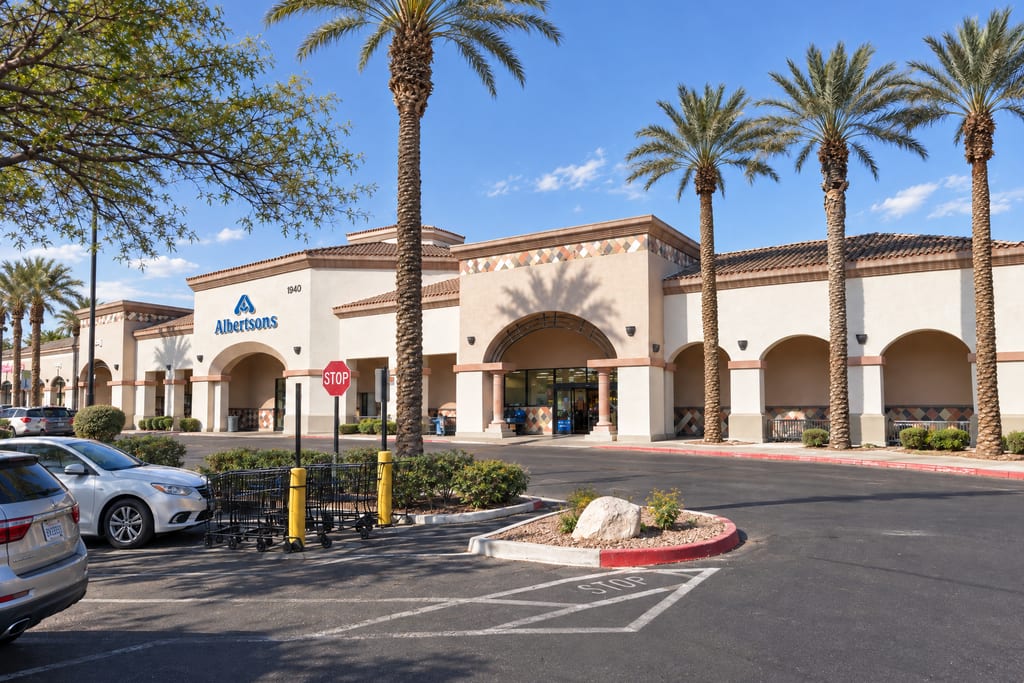 An Albertsons store numbered 1940 in a stucco shopping center with tile roofs and arched walkways, fronted by tall palms, a row of shopping carts and parked cars.