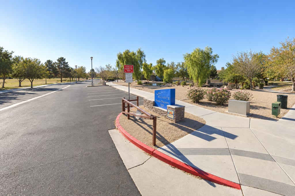 A blue Aliante Nature Discovery Park sign on a low stone base sits at the corner of a parking lot, with a no-feeding-the-birds sign beside it and pink oleanders, a pond and a small rock waterfall under willow trees behind it.