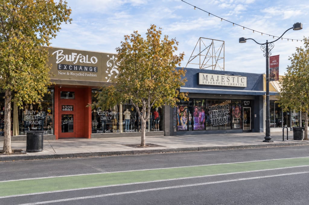 Buffalo Exchange and the Majestic Repertory Theatre side by side on a tree-lined sidewalk, with an Arts District banner on a lamppost, string lights overhead and a green bike lane in the street.