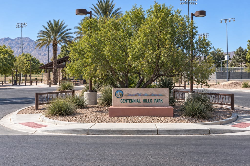 A stone Centennial Hills Park sign in a planted median, with palms, a picnic shelter, sports-field lights and desert mountains behind it.