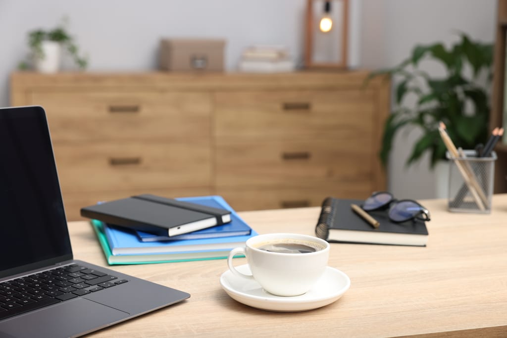 A laptop, coffee cup, notebooks and glasses on a home desk