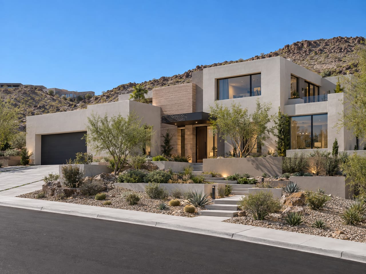 A contemporary stucco home with a layered tan accent wall, warmly lit windows and terraced desert planting and steps, against a rocky hillside under a clear sky