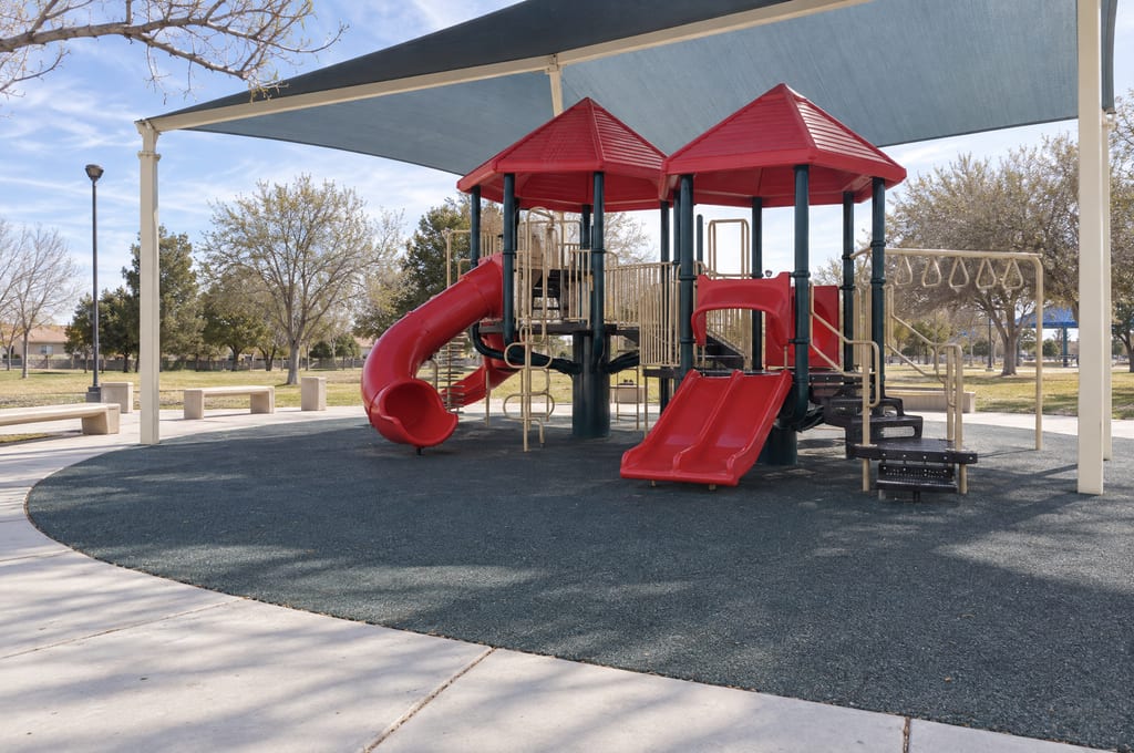 A red-roofed play structure with a tube slide and a double slide, under a large shade sail on rubber surfacing, with benches and a lawn of leafless trees behind it.