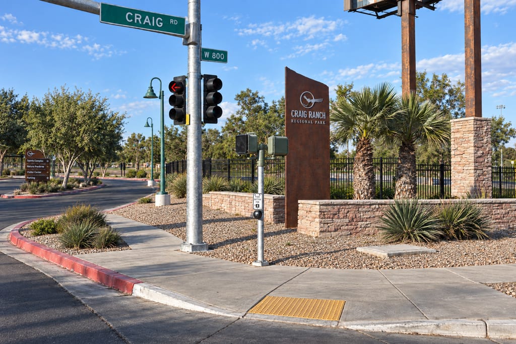 A rust-colored Craig Ranch Regional Park monument stands behind a stone planter wall at a corner with a traffic signal and a Craig street sign, with palms, an iron fence and park trees beyond.