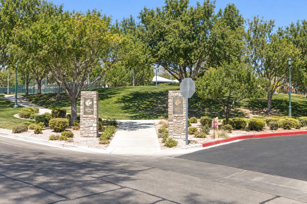 Two stone pillars with The Gardens Park plaques frame a walkway that climbs a grassy, tree-shaded slope toward a white shade canopy, with fenced courts at the left.