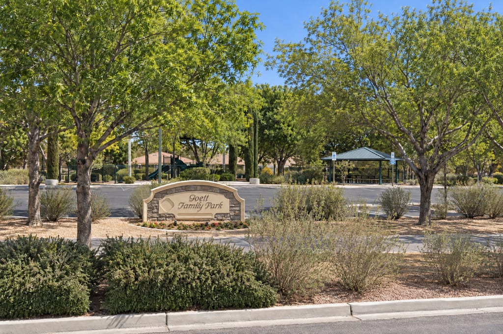 A stone monument sign reading Goett Family Park stands in a planted median, with shade trees, a fenced playground and a green-roofed picnic shelter across the parking lot behind it.