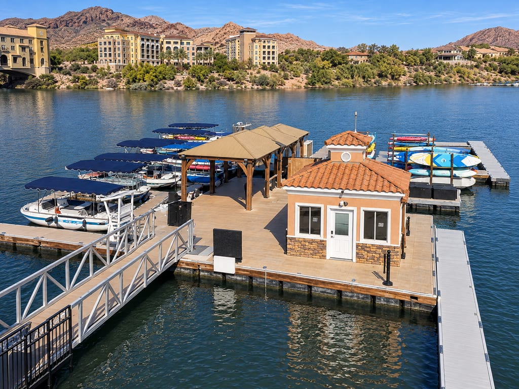 A floating dock with a small tile-roofed hut, a wooden shade pavilion, canopied boats and racks of kayaks, with hotels, hillside homes and mountains across the lake.