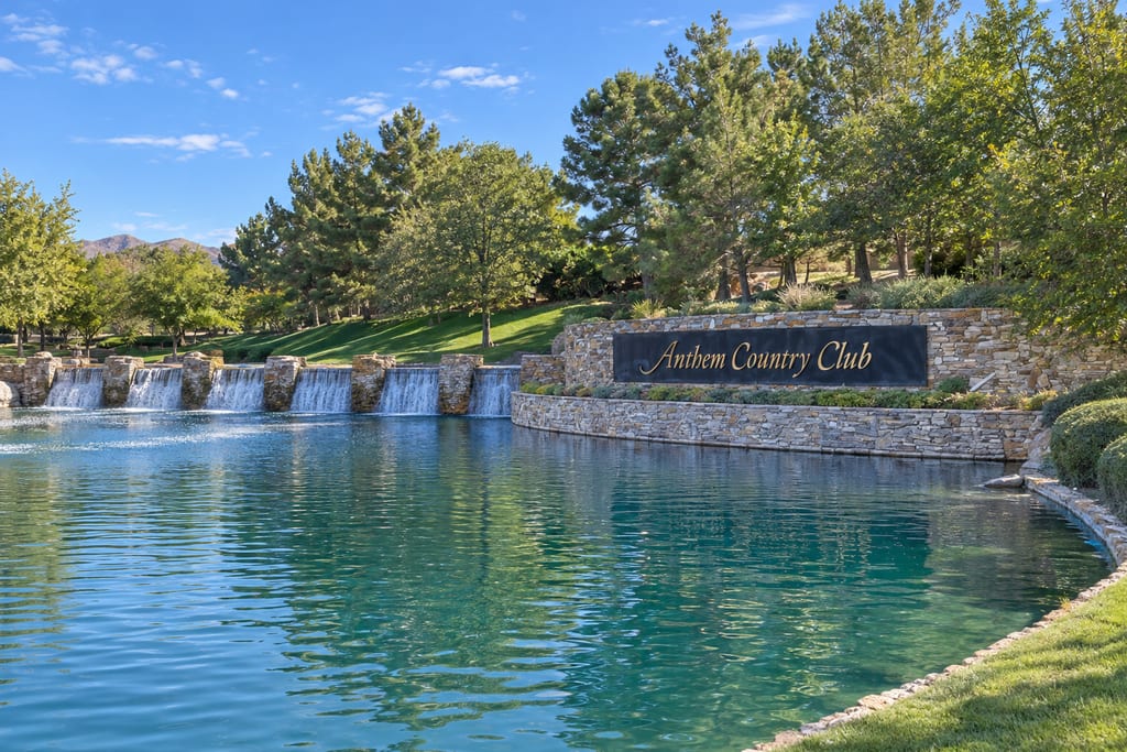 A stone wall with a black “Anthem Country Club” sign beside a pond with a row of small waterfalls