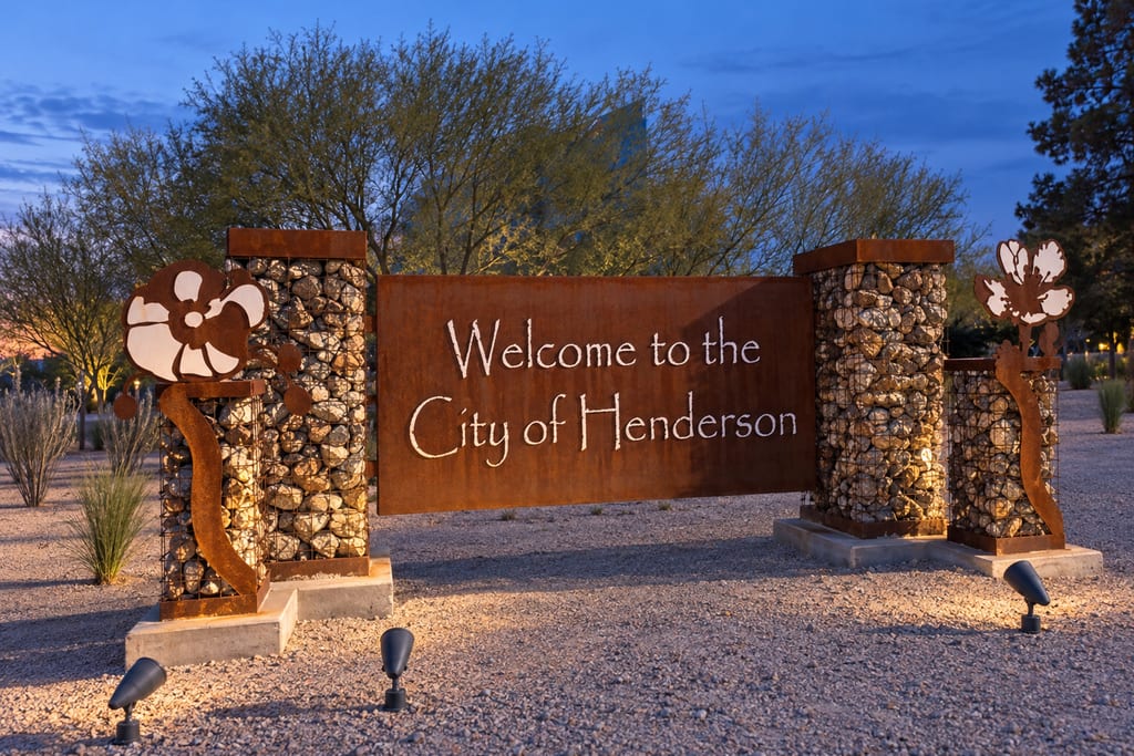 A rusted-steel sign reading “Welcome to the City of Henderson” between two stone-filled pillars, lit at dusk