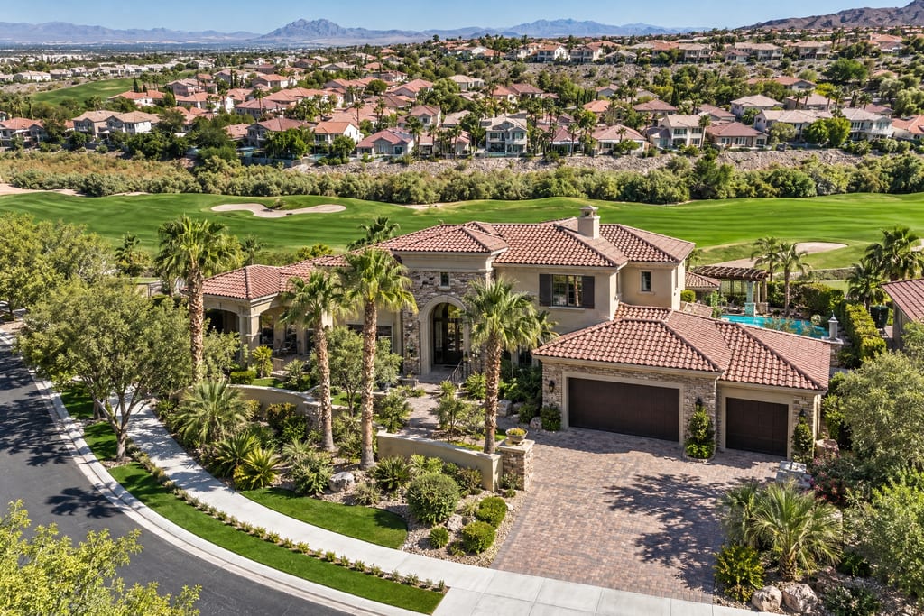 Aerial view of a large tile-roofed house beside a golf course, with more homes on the hillside behind it and mountains in the distance