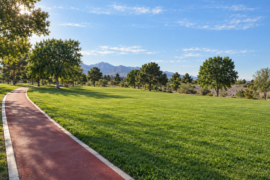 A red walking path beside a wide lawn and shade trees, with mountains in the distance
