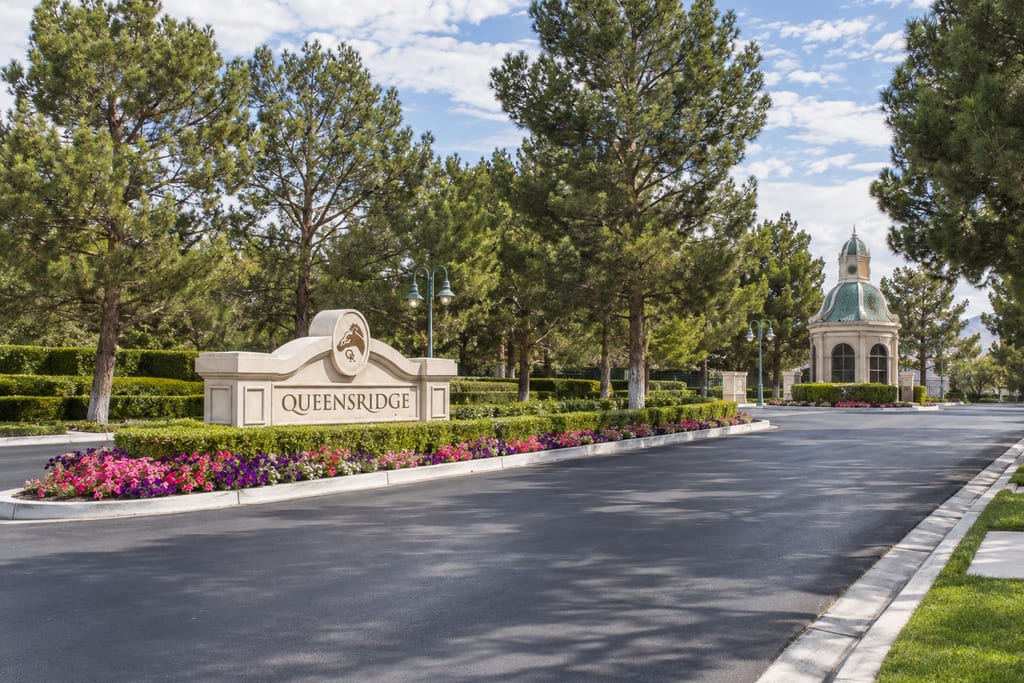 A cream stone entrance sign reading “Queensridge” among clipped hedges and flowers, beside a wide entry road