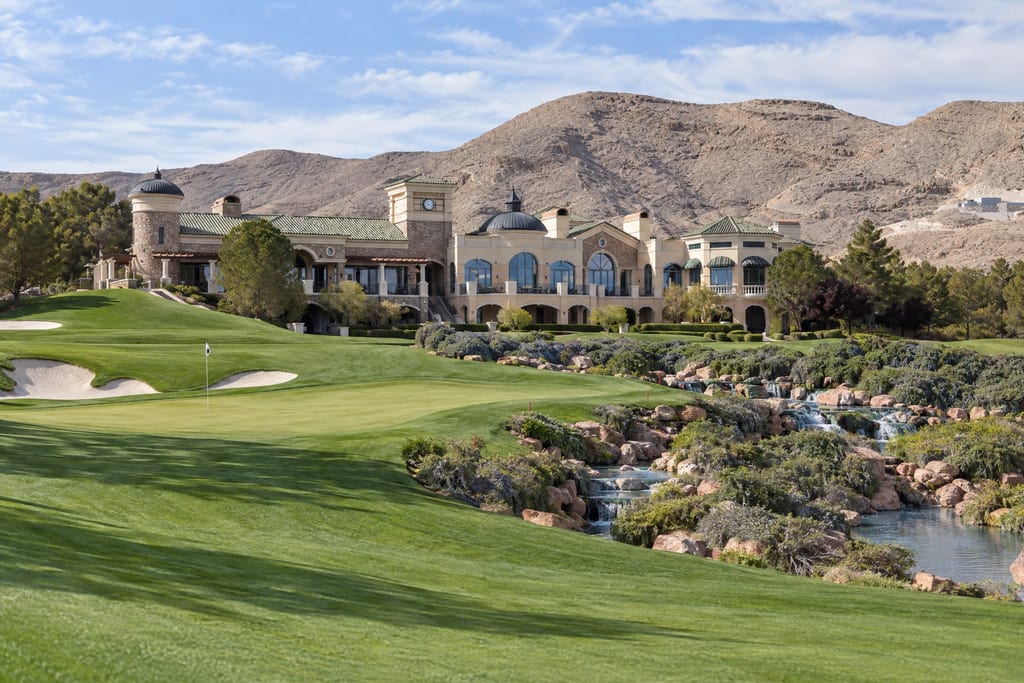 A large cream-stone clubhouse with a domed tower above a golf course and a rocky stream, bare mountains behind