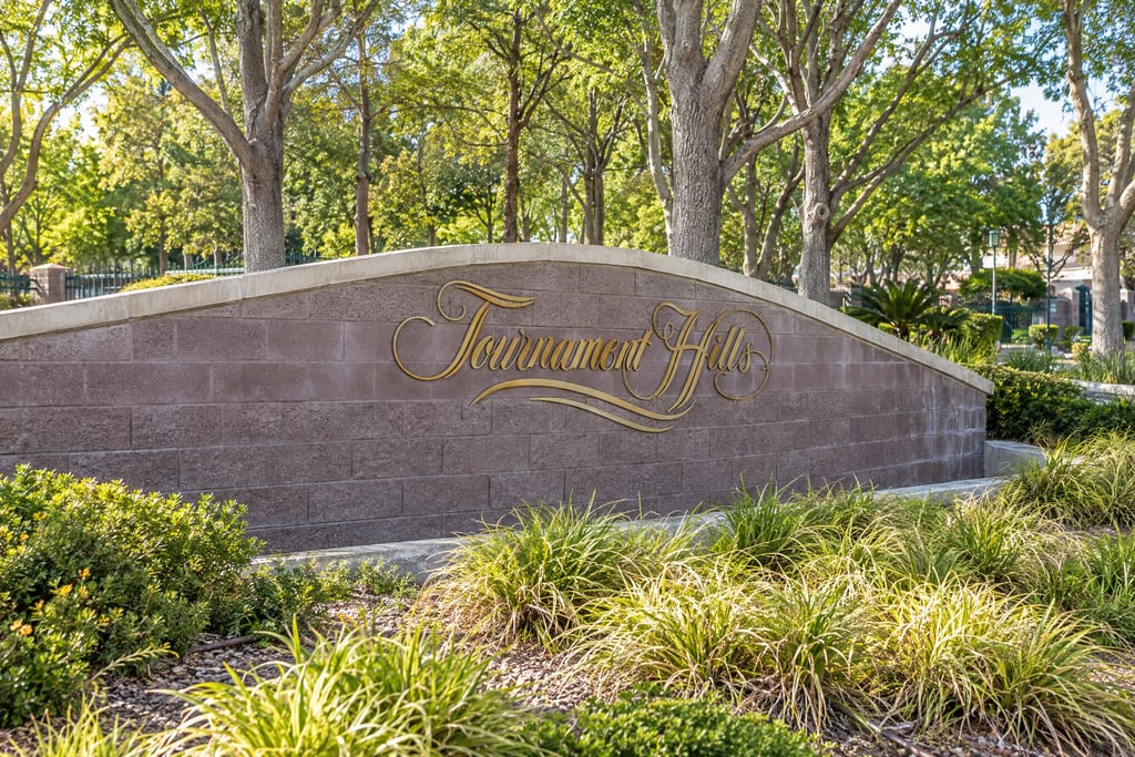 A curved entrance wall with “Tournament Hills” in gold script, shaded by trees