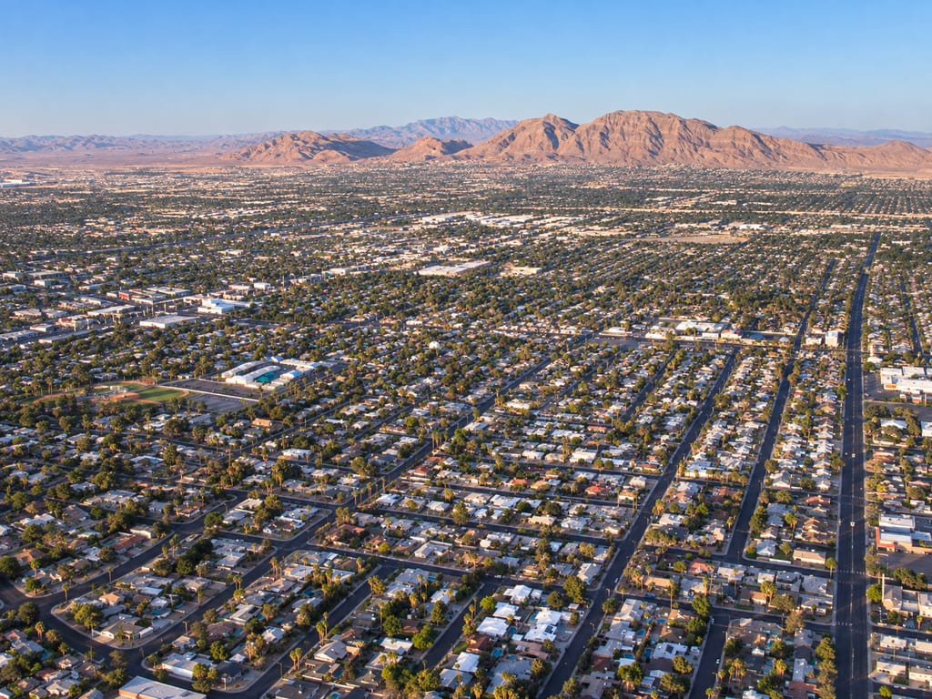 Rows of single-story homes with trees and backyard pools fill a grid of streets that runs toward bare mountains, with a ballfield at the left.