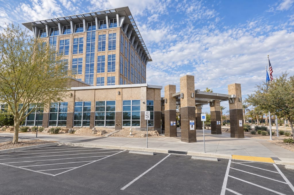 A tall tan stone and glass tower with a flared metal sunshade at its top rises behind a low wing and a pillared entrance canopy, with American and Nevada flags at the right and a parking lot in front.