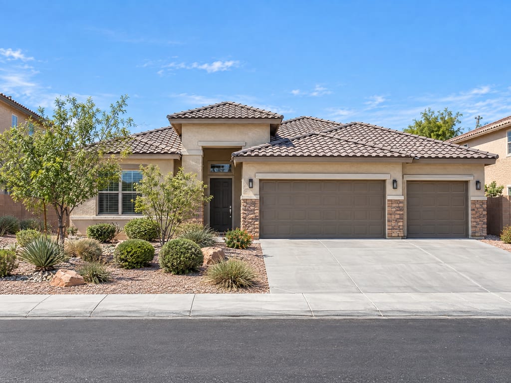 A single-story tan stucco house with a tile roof, stone trim, a double and a single garage door, and a front yard of desert plants and boulders.