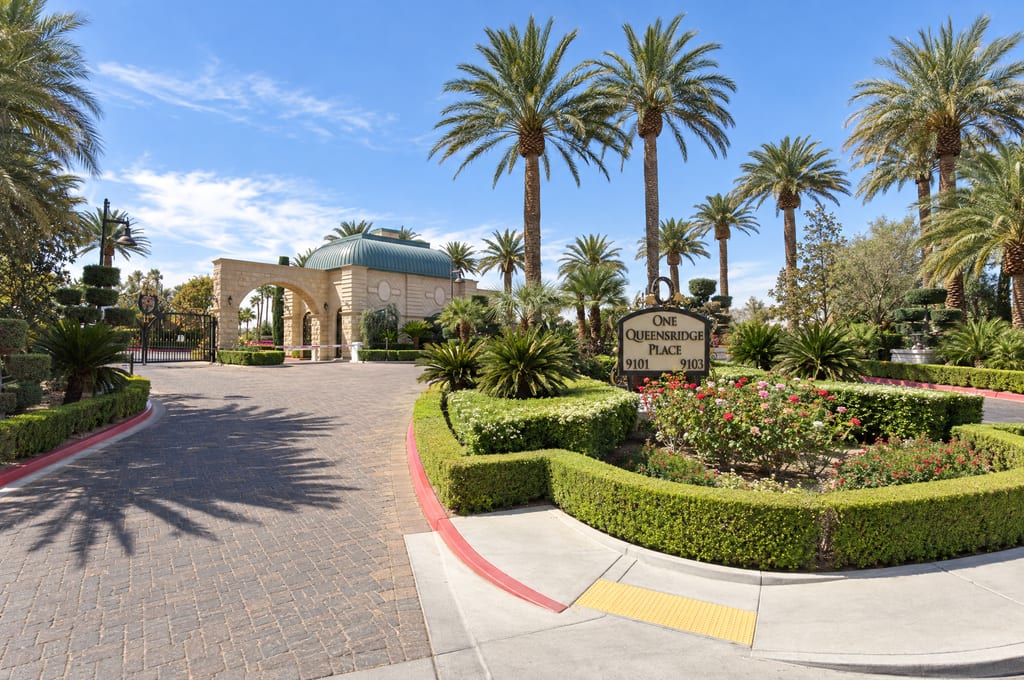 A sign reading One Queensridge Place, 9101 and 9103, stands in a bed of roses and clipped hedges beside a brick-paved drive that runs under tall palms to a domed stone gatehouse arch.