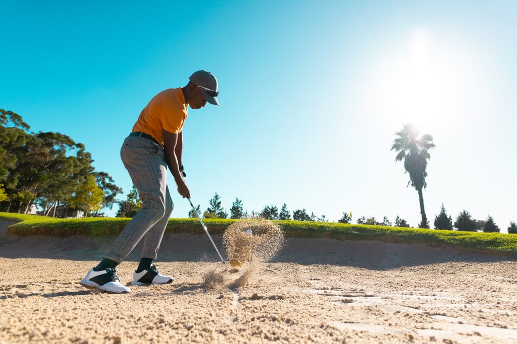 A golfer playing a bunker shot as the ball and sand rise into the sunlight