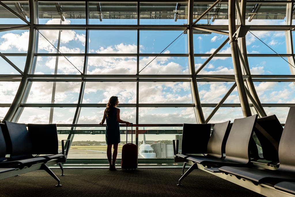 A traveler with a rolling suitcase beside tall airport windows overlooking a parked aircraft