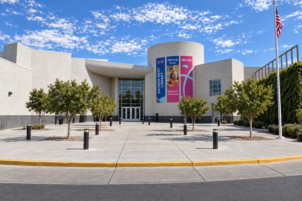 Sahara West Library banners hang on the round tower of a pale concrete building next to a tall glass entrance, at the end of a wide plaza with young trees, black bollards and a flagpole flying the American flag.