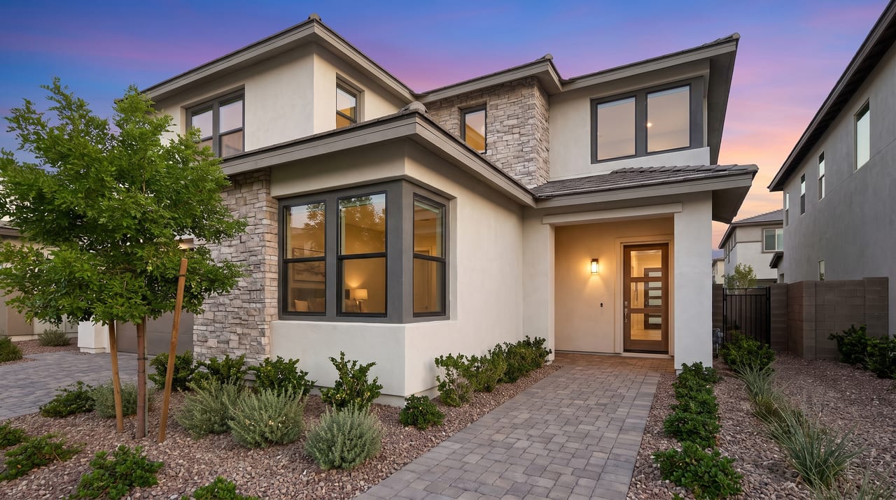 The lit front entry of a two-story stucco and stone home at dusk, with a paver walkway and desert plants