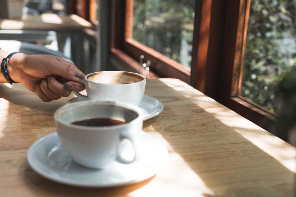 Two cups of coffee on a sunlit table beside a window.