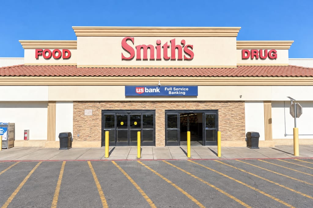 The entrance of a Smith's Food and Drug store, a stone-clad wall with two sets of glass doors under a red tile roof and a U.S. Bank Full Service Banking sign, behind yellow posts and an empty parking lot.