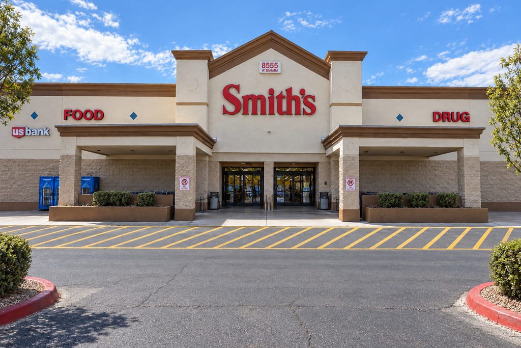 The entrance of a Smith’s Food and Drug store under the address 8555 W. Sahara, a peaked stucco front between two covered walkways, facing a striped fire lane and an empty parking lot.