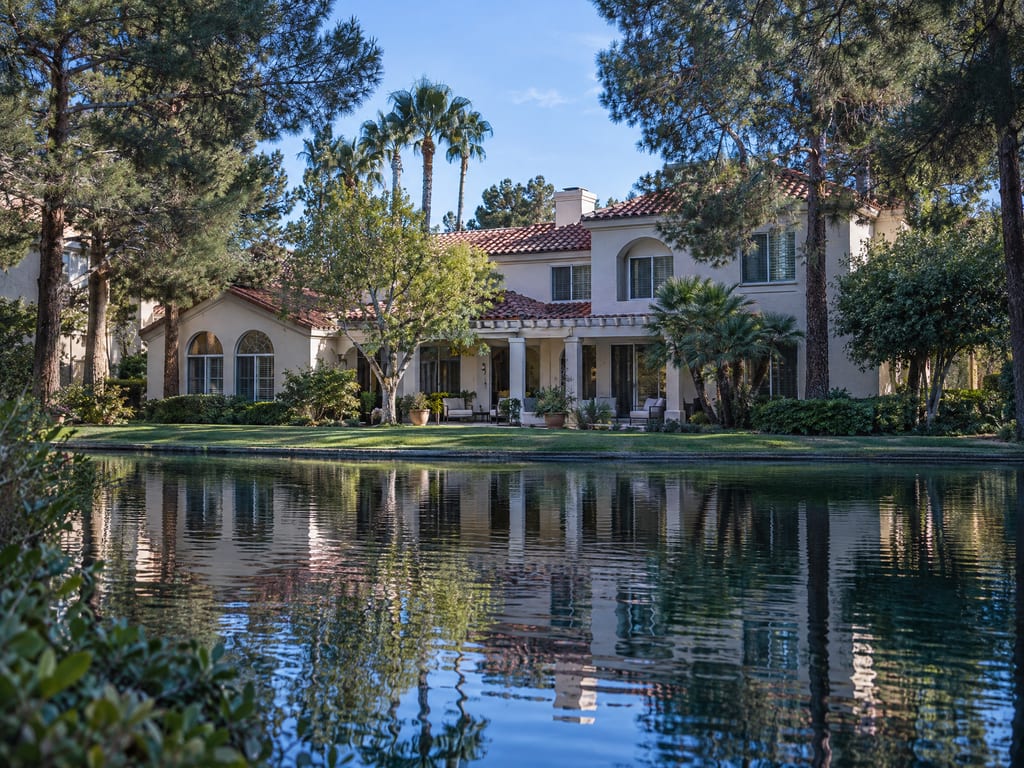 A two-story stucco house with a red tile roof and a columned patio sits on a lawn across calm water, framed by pines and palms.