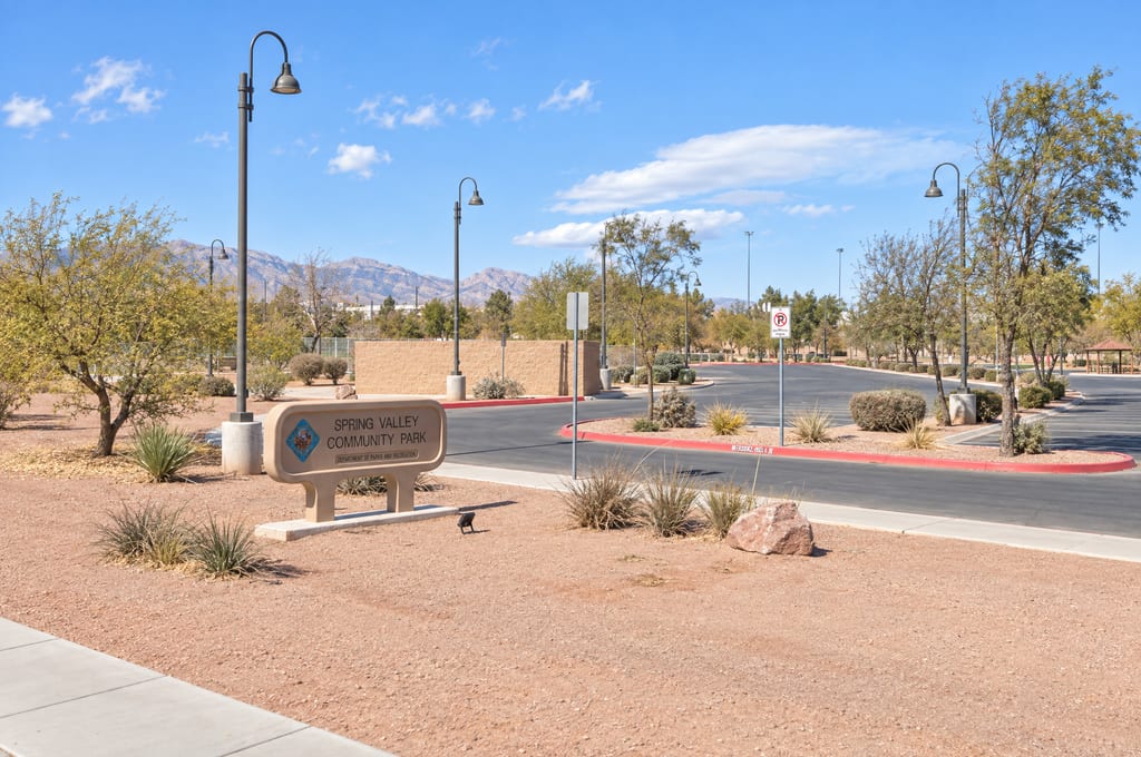 A sign reading Spring Valley Community Park, Department of Parks and Recreation, stands in a gravel bed beside the park's entrance drive, with lamp posts, a block wall and trees in front of a mountain range.