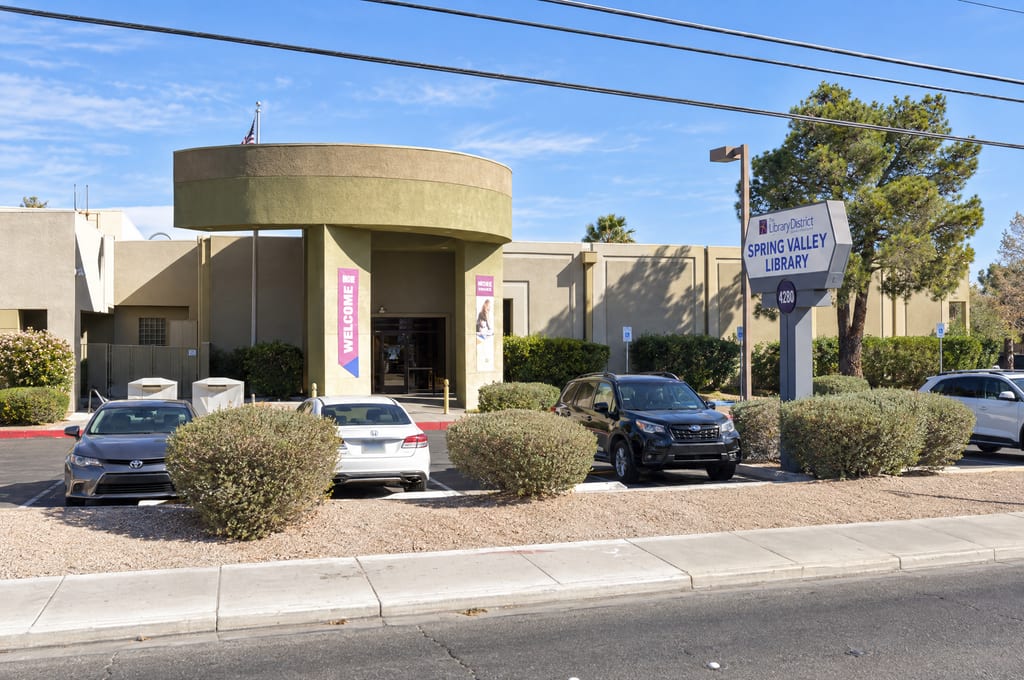 A Library District sign reading Spring Valley Library above the number 4280 stands beside the parking lot of a tan stucco library with a round entrance canopy, pink welcome banners and a flag on the roof.