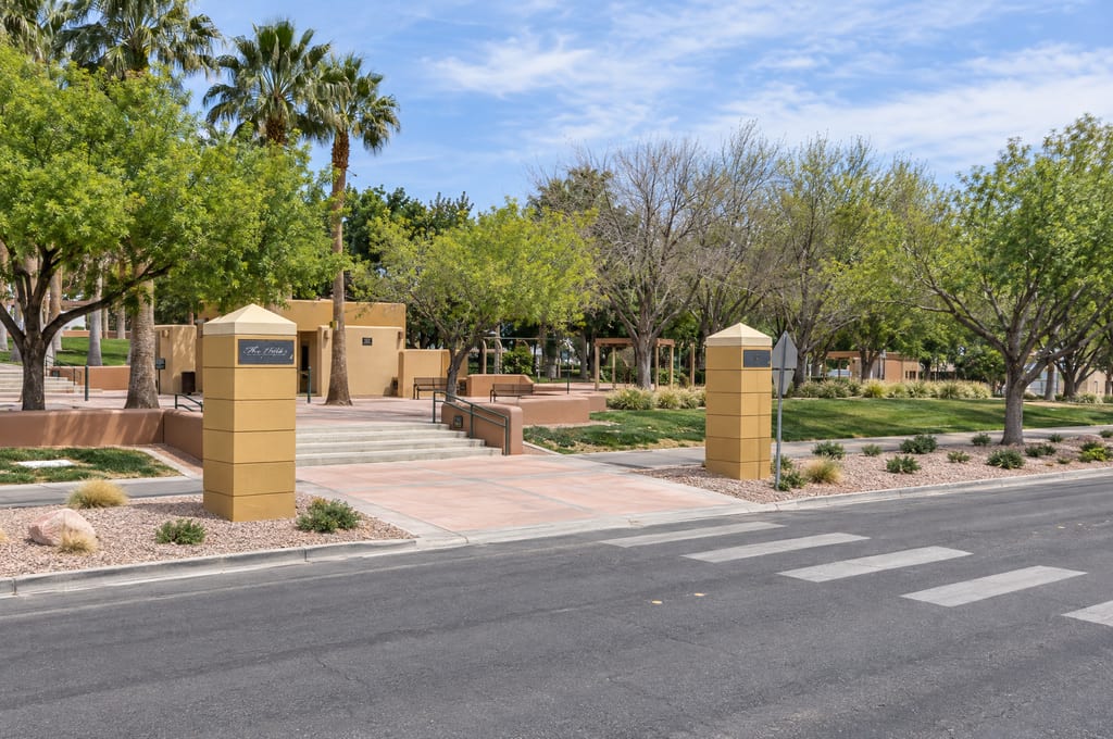 Two tan stucco pillars, one with a plaque reading The Hills, frame a crosswalk entrance with steps up to a small adobe-style building, benches and a shade pergola under palms and leafy trees.