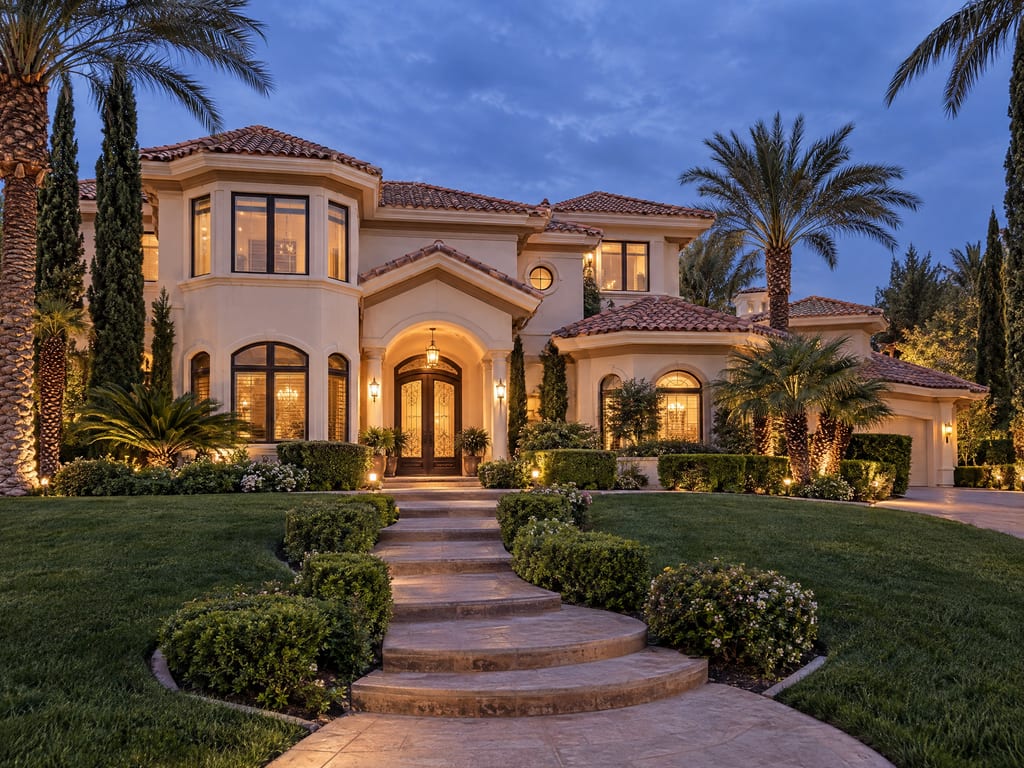 A two-story cream stucco house with red tile roofs and lit windows at dusk, with curved stone steps climbing through a lawn to an arched front door.