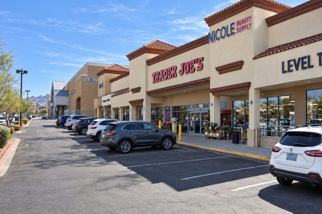 A tan strip center with tile-roofed towers has a red Trader Joe’s sign over glass doors marked 180, with potted flowers by the entrance and cars parked along the lot.