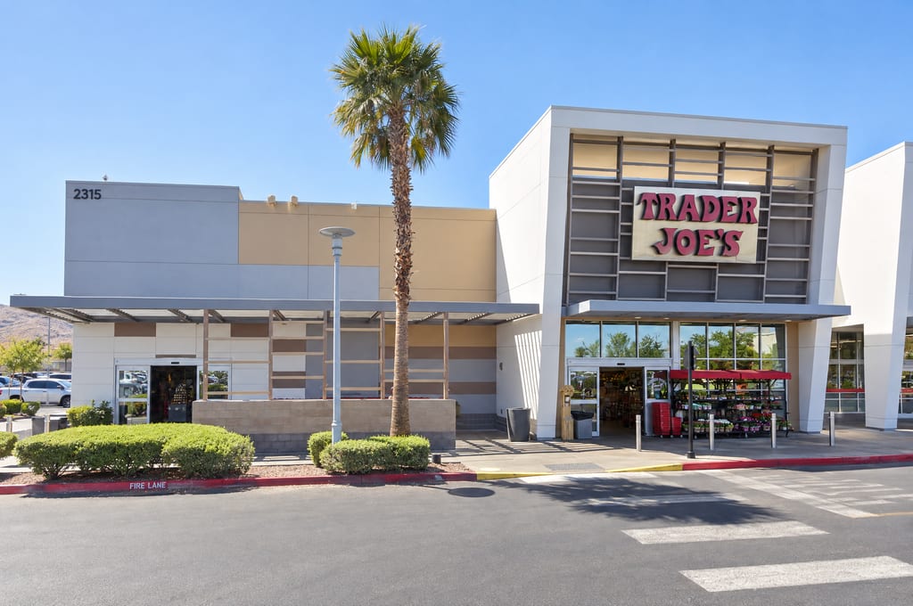 A white and gray Trader Joe's with its red sign above glass doors and a flower stand, next to a covered side entrance under the number 2315, a tall palm and a crosswalk across the fire lane.