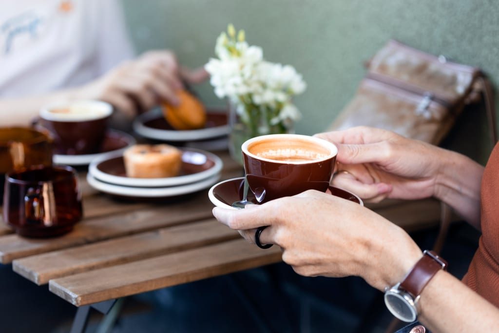 Hands holding a cup of coffee beside cake and flowers on a café table