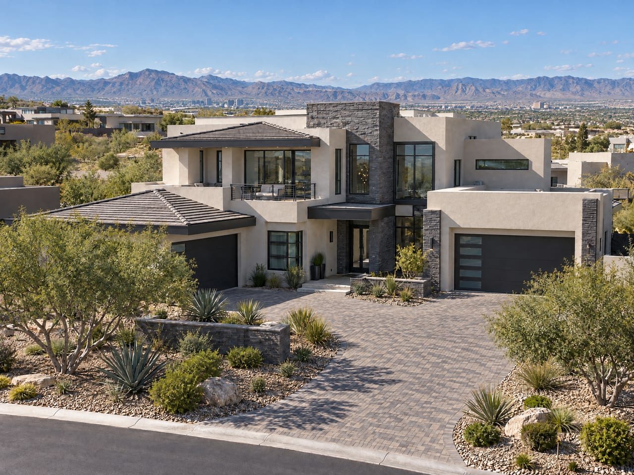 A contemporary two-story stucco and stacked-stone home with a paver driveway and desert landscaping, seen from above, with the valley, a distant skyline and mountains beyond