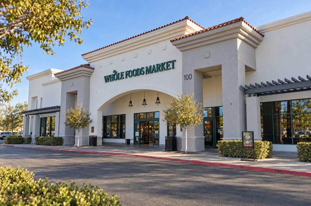 The arched entrance of a Whole Foods Market, a cream stucco building with a red tile roof and the street number 100 on its pillar, with young trees along the sidewalk and an empty drive in front.