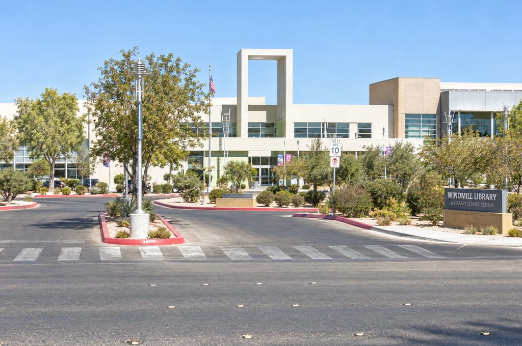 The Windmill Library sign beside the entrance drive, with the pale multistory library and a tall open rectangular tower behind a row of trees.